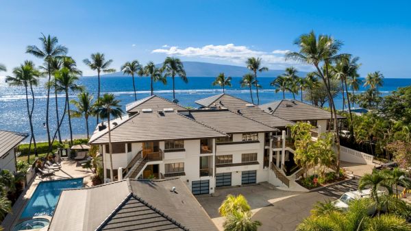 Tropical resort with brown-roofed villas, palm trees, a swimming pool, and the ocean in the background under a bright blue sky.