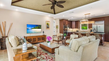 Spacious, bright living room with beige sofas, wooden coffee table, tropical plants, wall-mounted TV, and an open kitchen with dark wood cabinets and a island, ceiling fan, and tiled floor.