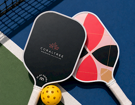 Two beach tennis paddles and a yellow perforated ball on a blue-green court with a net in the background.
