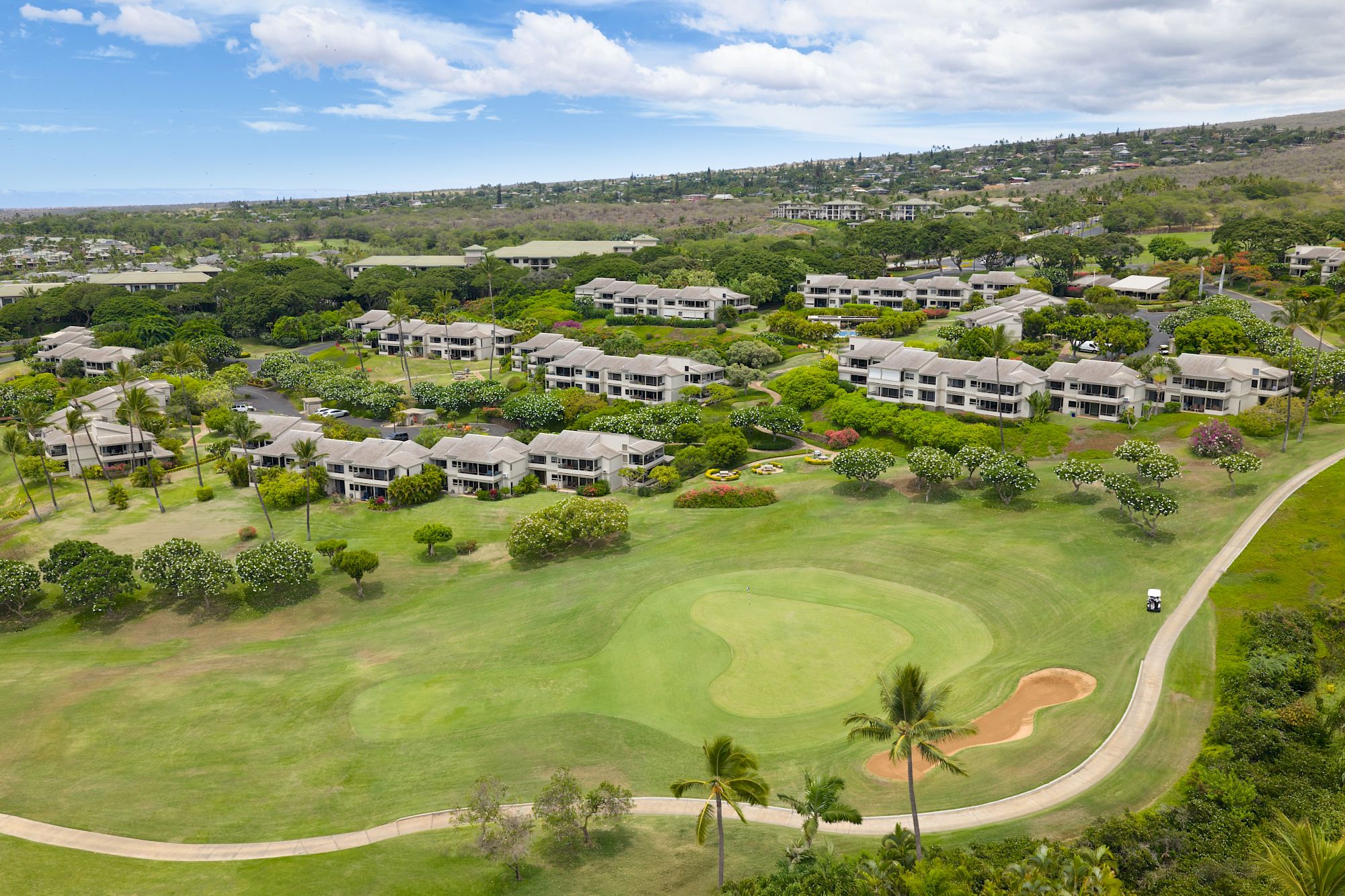 Aerial view of a resort with light-colored buildings around a lush green golf course, palm trees, and a winding road through a sunny, tropical landscape.