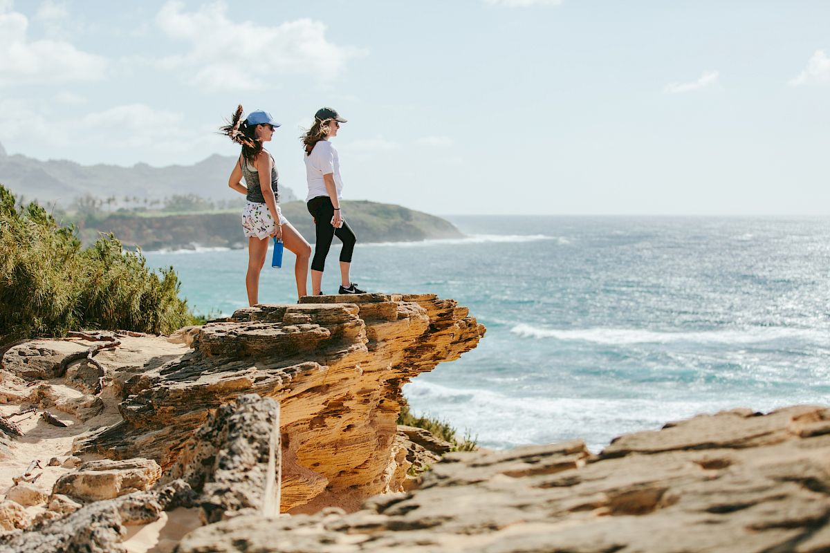 Two people stand on a rocky cliff above the ocean, gazing at the horizon as waves crash below on a sunny day.