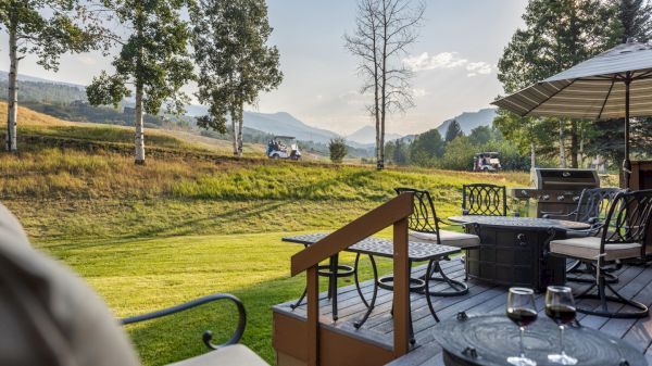 A serene outdoor patio overlooking a lush lawn and distant mountains, with chairs, tables, and a few trees under a sunny sky.