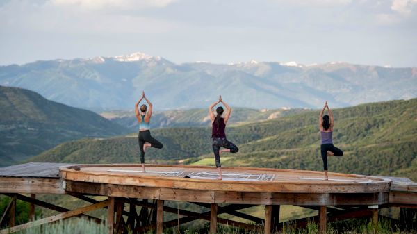 Three people are practicing yoga on a wooden platform with a mountain landscape behind, balancing in tree pose.