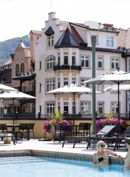 A European-style hotel with pastel facades, outdoor pool, sun loungers and two people swimming.