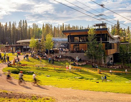 A modern lodge perched by a sunny lawn, people enjoying outdoor seating, pathways, and a small stage near a forest backdrop.