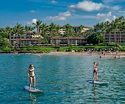 Two people stand up paddling on a sunny beach with clear blue water, a tropical resort lined with palm trees in the background.