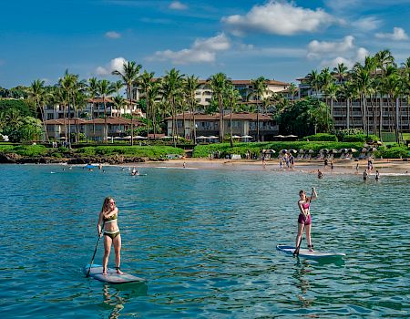 Two people stand up paddling on a sunny beach with clear blue water, a tropical resort lined with palm trees in the background.