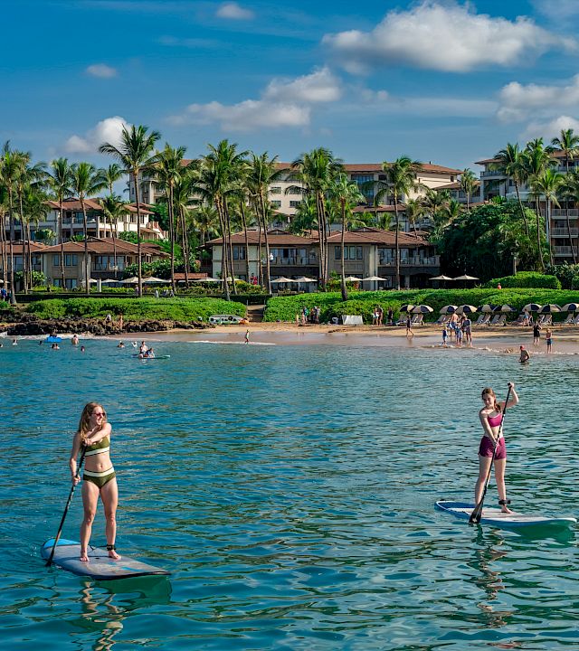 Two people stand up paddling on a sunny beach with clear blue water, a tropical resort lined with palm trees in the background.