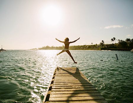 A person jumps joyfully from a dock into a sunny, calm lake with a bright horizon and a beach shoreline in the background.