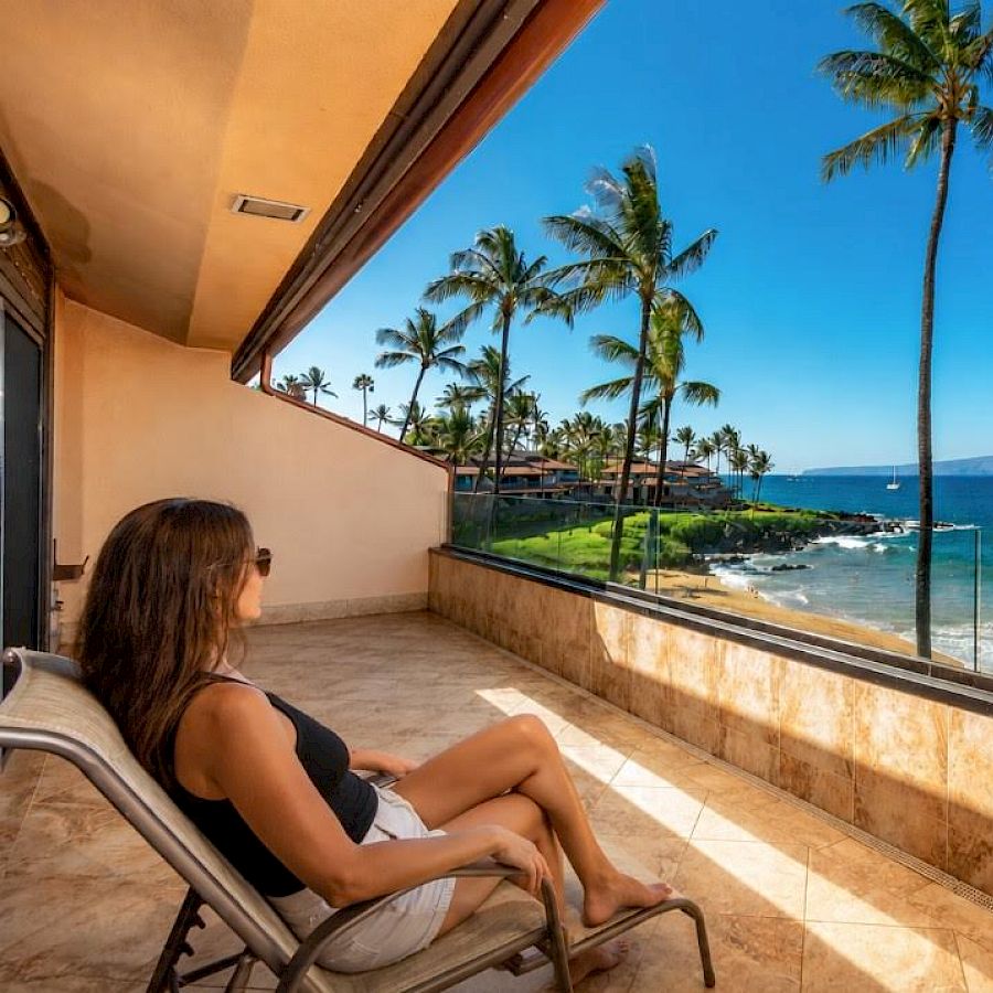 A person lounges on a patio chair, overlooking a tropical beach with palm trees, turquoise waves, and clear blue skies beyond a glass railing.