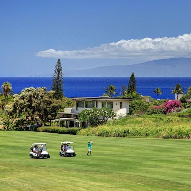 Two golf carts on a manicured green, a person walking, with stylish houses and lush tropical plants, and a bright blue ocean beyond.