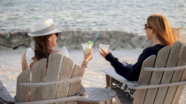 Two women relax on wooden chairs by the beach, sipping cocktails and chatting as waves roll in at sunset.