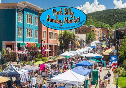 A lively outdoor market scene with colorful tents, shoppers, and a blue sign reading &ldquo;Park Silly Sunday Market&rdquo; over a sunny mountain town street.