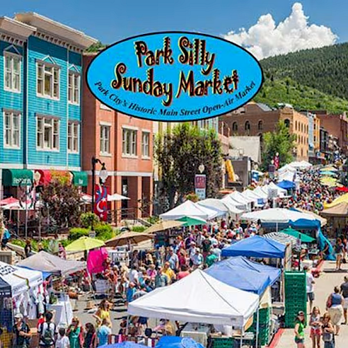 A lively outdoor market scene with colorful tents, shoppers, and a blue sign reading “Park Silly Sunday Market” over a sunny mountain town street.