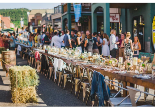 A long rustic street feast with a row of tables set for a crowd, hay bales, string lights, and people mingling along a sunny outdoor market scene.