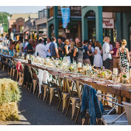 A long rustic street feast with a row of tables set for a crowd, hay bales, string lights, and people mingling along a sunny outdoor market scene.