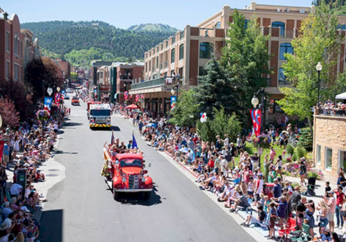 A street parade with a crowd on both sides watching red convertibles drive by, lined by buildings and mountains in the background.