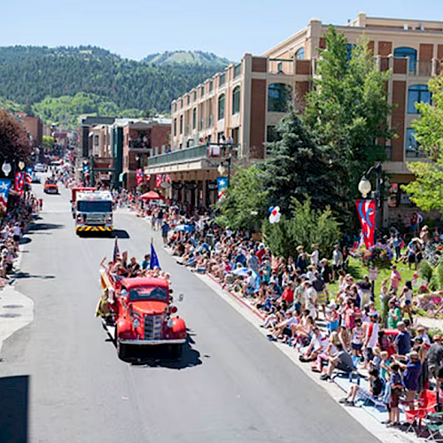 A street parade with a crowd on both sides watching red convertibles drive by, lined by buildings and mountains in the background.