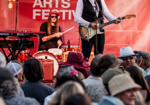 A live band performs on stage at Kimball Arts Festival, with a guitarist and drummer in front of a cheering crowd.