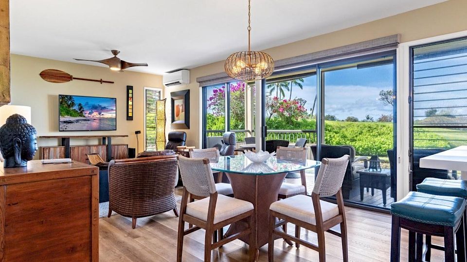 A bright, open living-dining area with a round table, six chairs, a ceiling fan, large glass doors to a patio, and ocean view beyond.