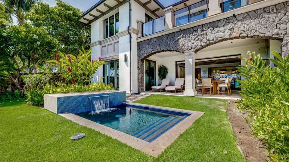 A modern two-story villa with a stone balcony, a small pool, and a covered patio overlooking a green lawn.