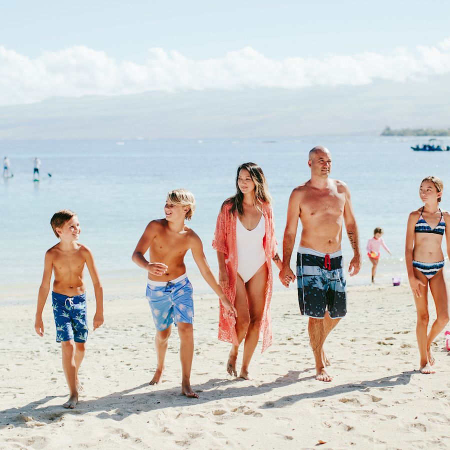A family walks along a sunny beach in swimsuits, enjoying the calm sea and clear skies, with boats in the distance and footprints in the sand.