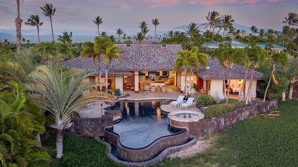 A tropical villa with a curved pool, palm trees, and a sunset backdrop, surrounded by lush greenery and lounge chairs, exuding luxury.