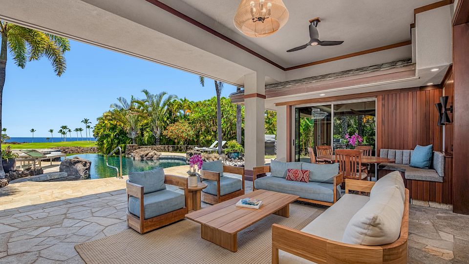 Cozy outdoor living area on a sunny patio with wooden furniture, neutral cushions, and a view of a lush garden, pool, and ocean beyond.