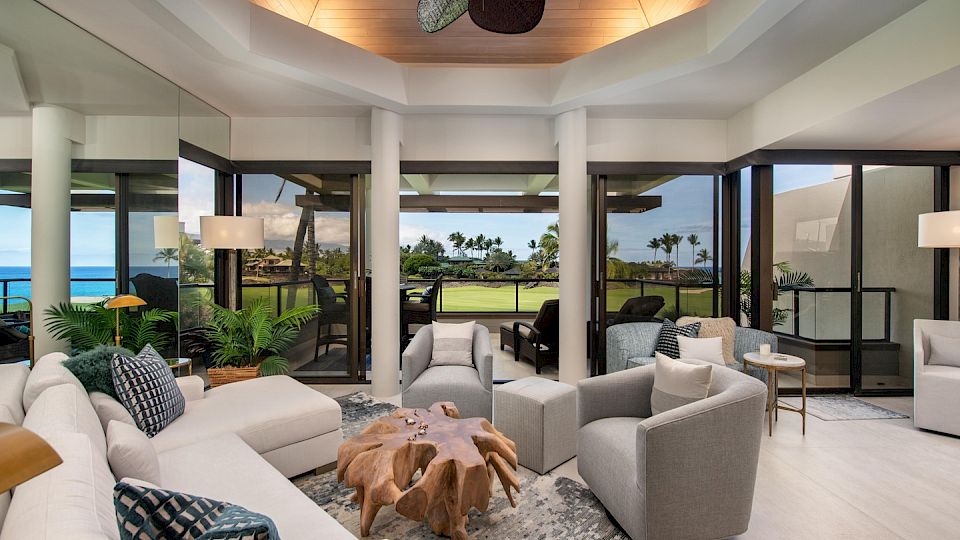 Luxurious, bright living room with white and gray seating, glass walls, tropical view, modern coffee table, ceiling fan, and open balcony.