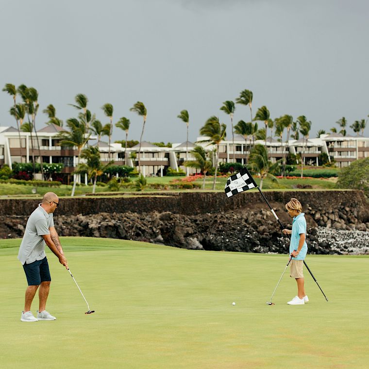 Two people playing golf on a sunny, grassy course near the coast, with palm trees and buildings in the background, a flag near the hole.
