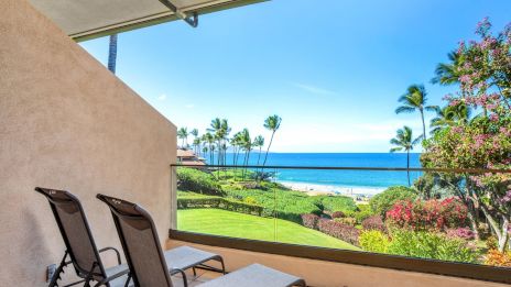A balcony with two lounge chairs overlooks a tropical lawn, palm trees, and a blue ocean under a clear sky. END.