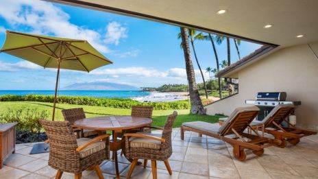 A tropical patio with a round wicker table, four chairs, a green umbrella, lounge chairs, a grill, palm trees, and a view of the blue ocean and sky beyond.