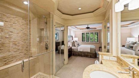 A luxurious hotel-style bedroom suite with a large glass-enclosed shower, double vanity, and a cozy bed area visible through an arched entryway.