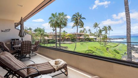 Luxurious balcony with loungers, a shaded seating area, and a view of palm trees, a grassy yard, and the ocean beyond under blue skies.