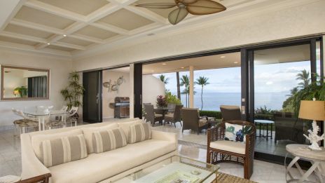 A bright, airy living room with beige sofa, glass coffee table, indoor plants, and sliding glass doors opening to a ocean-view patio.