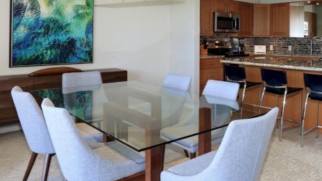 A modern dining area with a glass-top table, six gray upholstered chairs, and a kitchen with wooden cabinets and a breakfast bar.