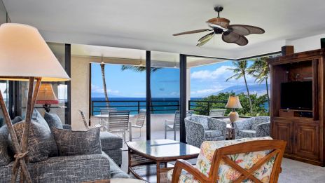 A bright living room with wicker chairs and sofas, facing a glass wall to a balcony that overlooks blue ocean water and palm trees outside.
