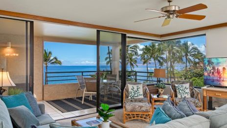 A bright coastal living room with a ceiling fan, ceiling-to-floor windows, palm trees outside, and a view of the ocean.