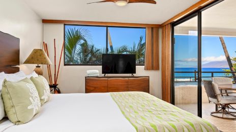A bright hotel room with a large bed, green accents, a TV on a dresser, sliding glass doors to a balcony, and ocean view outside.