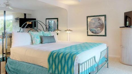 A bright, modern bedroom with a metal bed frame, white bedding, turquoise throw, matching pillows, a large mirror, framed wall art, and a curved dresser or cabinet in the corner.