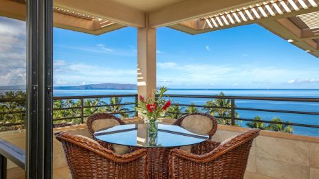 Ocean view balcony with a glass-top table, four wicker chairs, and a vase of red flowers; clear blue sky and calm sea beyond.