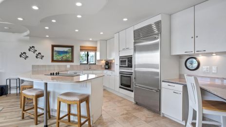 A bright, modern kitchen with white cabinets, stainless steel appliances, a center island with stools, and a built-in desk area. ends.
