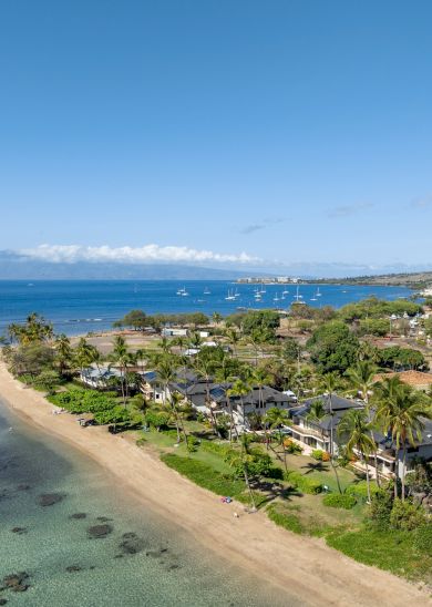 A sunny coastal scene with a sandy beach, turquoise water, and a small resort town of palm trees and low-rise buildings along the shore.