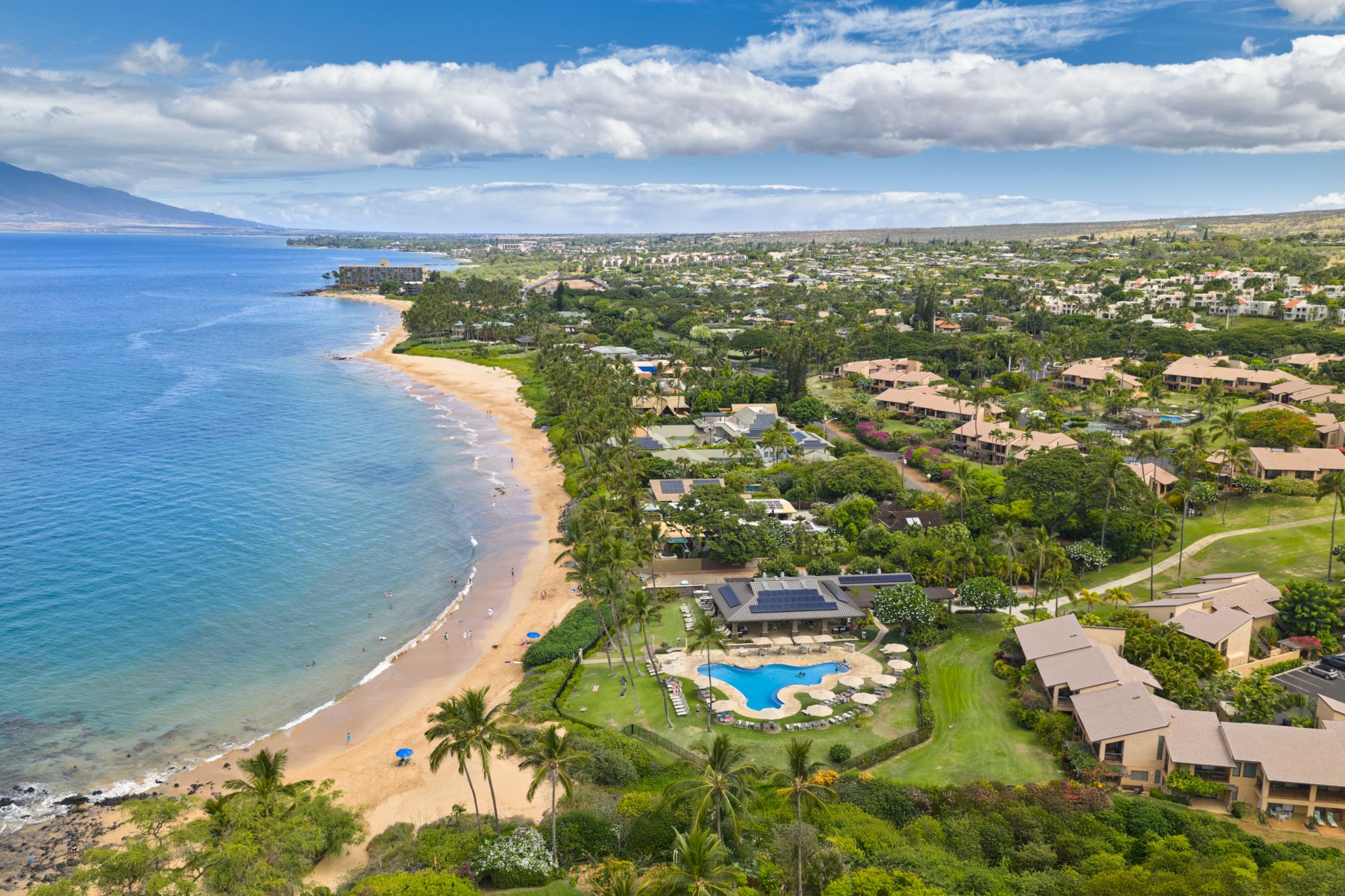 A tropical coastal community with a sandy beach, clear blue bay, lush greenery, and resort-style buildings including a pool, all along the shoreline. End.