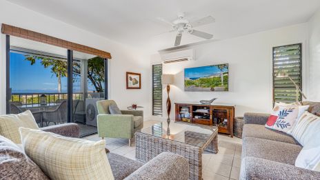 A bright, seaside living room with a wicker coffee table, gray sofas, a ceiling fan, a wall-mounted TV, and large sliding doors opening to a balcony with ocean views.