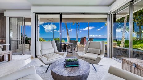 Luxurious indoor&ndash;outdoor living room opens to a sunny patio and ocean view, with comfy white chairs, a round table, and glass doors. end.