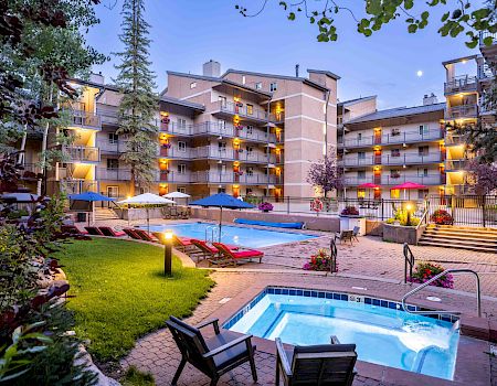 A sunny hotel courtyard with a central pool, surrounding balconies, lounge chairs, and a wading pool, nestled among trees and low-rise buildings.