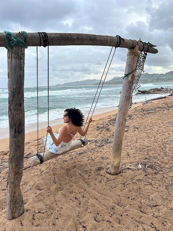 A woman in a white dress sits on a wooden swing on a sandy beach, waves and cloudy sky in the background.