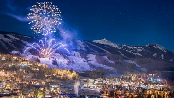 A snowy mountain town glows with festive lights as fireworks explode in a midnight sky above the rooftops.