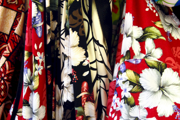 A row of colorful Hawaiian shirts with bold floral patterns&mdash;red, white, black, and blue hues, tropical leaves, and hibiscus designs.